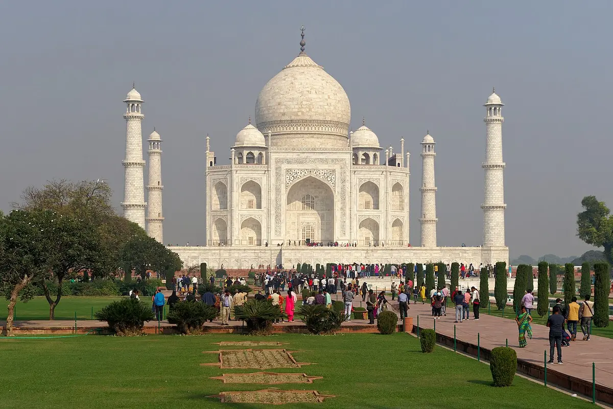 Crowds of visitors walking the garden paths toward the Taj Mahal under a hazy sky